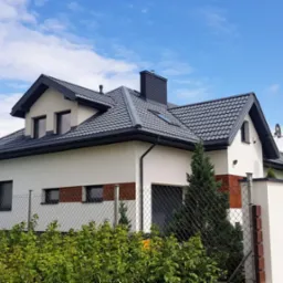 Nowoczesny dom jednorodzinny z ciemnoszarą dachówką ceramiczną, ciemnym orynnowaniem i kominem z cegły klinkierowej, widok zza ogrodzenia porośniętego zielenią.