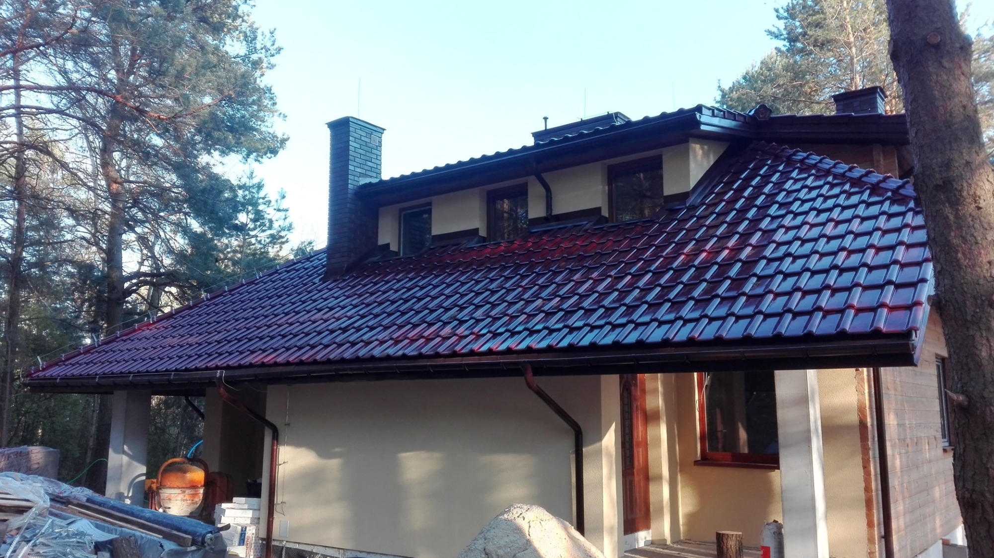 Dom jednorodzinny z nowym, błyszczącym, bordowym pokryciem dachowym w kształcie dachówki, z ciemnobrązowymi rynnami, widocznym kominem z cegły i fragmentem terenu budowy z betoniarką.