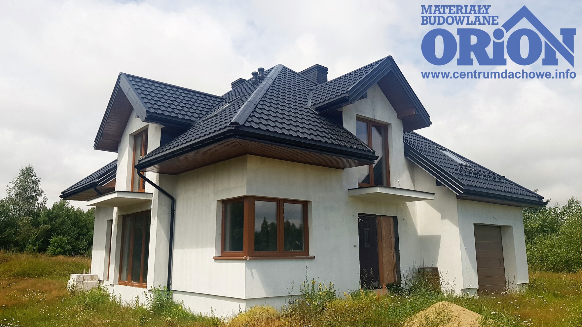 Nowy dom jednorodzinny z czarnym dachem z dachówki metalowej, brązowymi oknami i białymi ścianami zewnętrznymi, widoczne logo producenta na górze kadru.