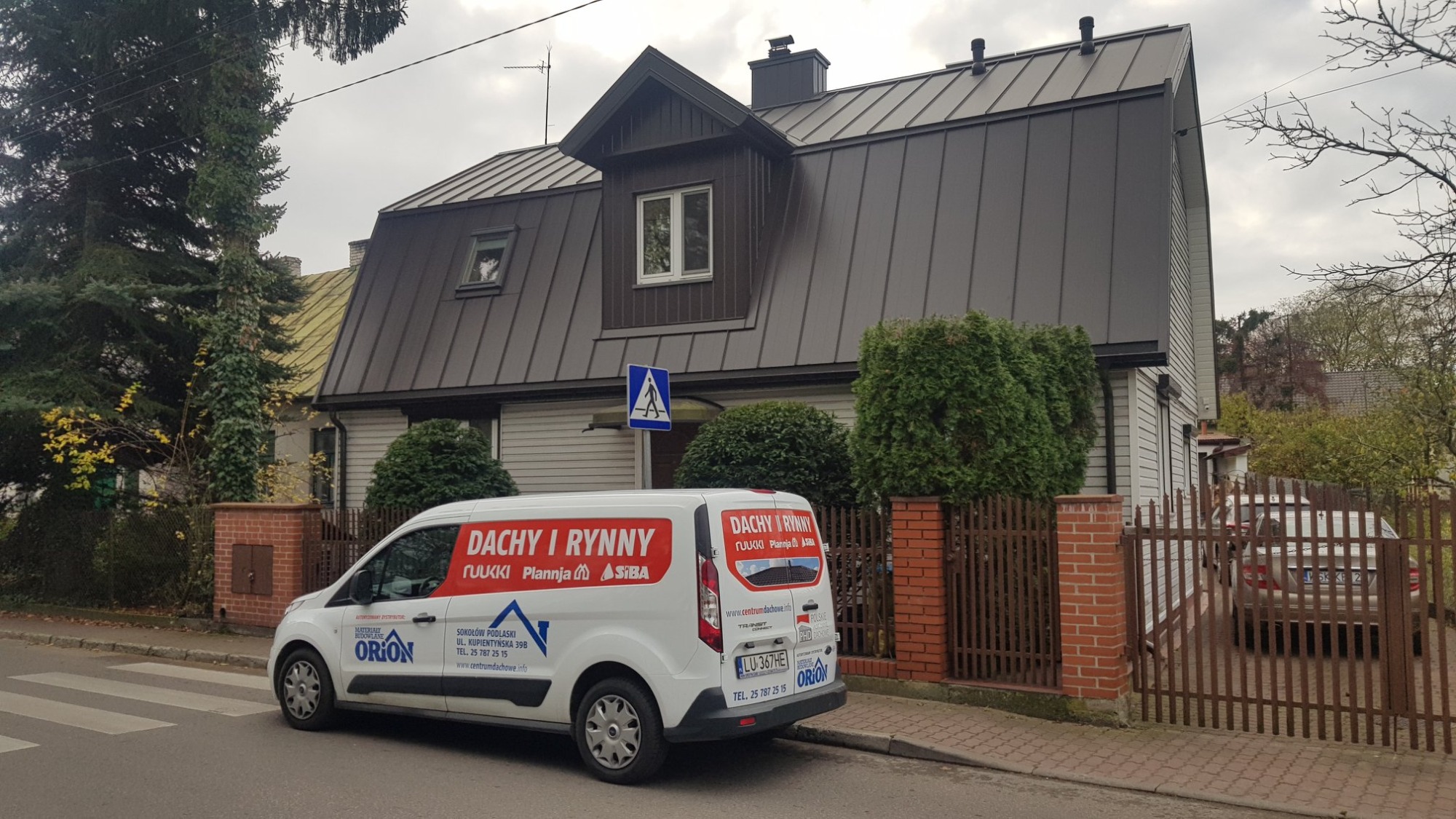 Dom jednorodzinny z nowym, ciemnym pokryciem dachowym, widoczny z ulicy, z zaparkowanym firmowym vanem z logo i informacjami kontaktowymi firmy dekarskiej.