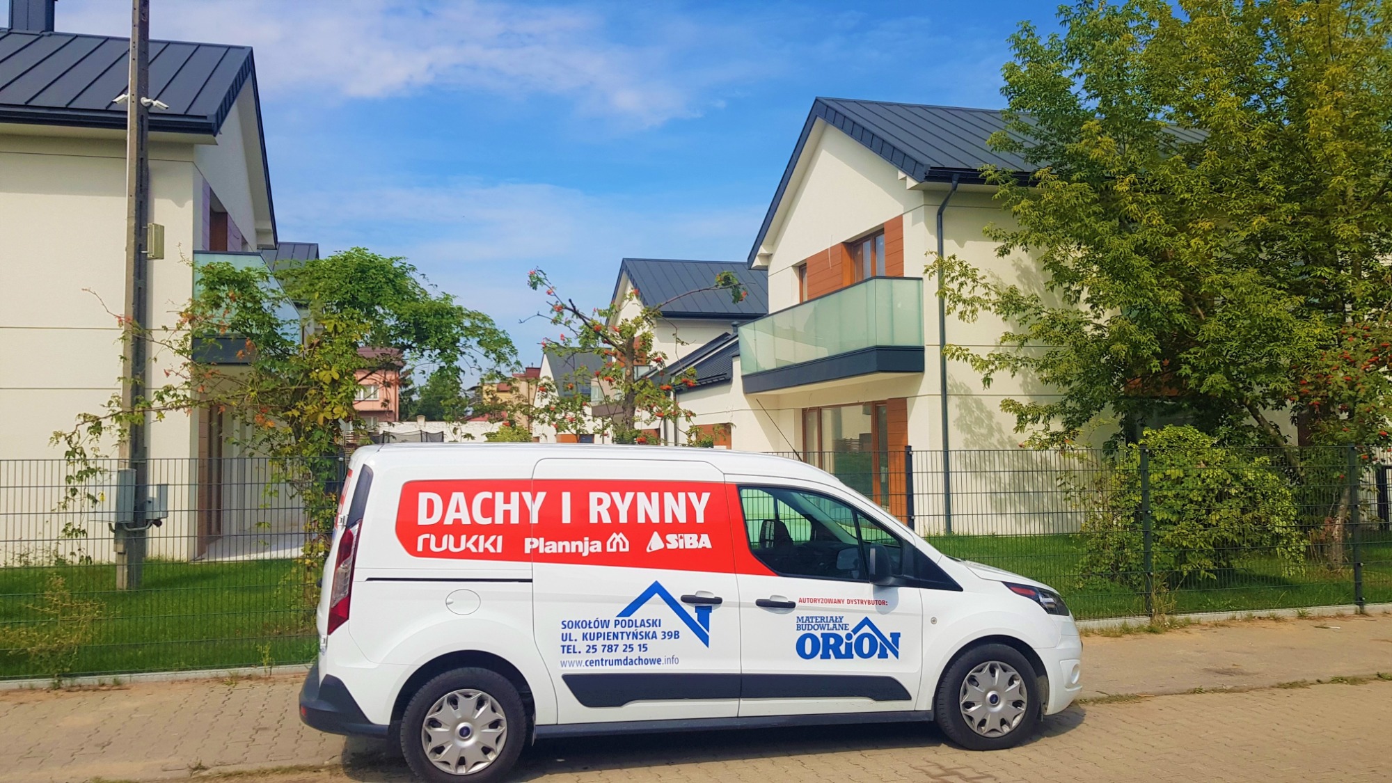 Biały samochód dostawczy z czerwonym nadrukiem 'Dachy i Rynny', zaparkowany na tle nowych domów z ciemnymi dachami i zielenią, prezentujący usługi dekarskie i dystrybucję materiałów budowlanych.