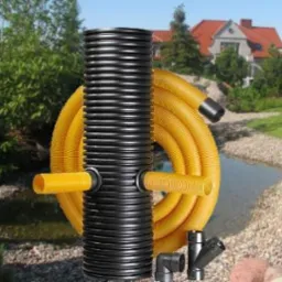 Czarna, karbowana kolumna drenażowa z żółtą perforowaną rurą drenażową zwiniętą obok, złączki kanalizacyjne na ziemi, w tle ogród z oczkiem wodnym i dom z czerwoną dachówką.