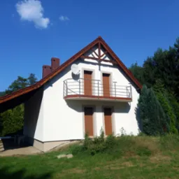 Biały dom jednorodzinny z brązowymi elementami wykończeniowymi, widoczna antena satelitarna na elewacji, otoczony zielenią i drzewami w słoneczny dzień.