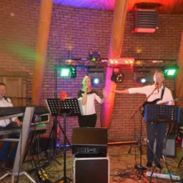 Zespół muzyczny na scenie z ceglanym tłem, oświetleniem scenicznym i instrumentami: keyboard Yamaha, gitara elektryczna. Wokalistka w centrum, mężczyźni po bokach.