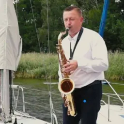 Saksofonista gra na saksofonie na pokładzie jachtu, w tle woda i zieleń.