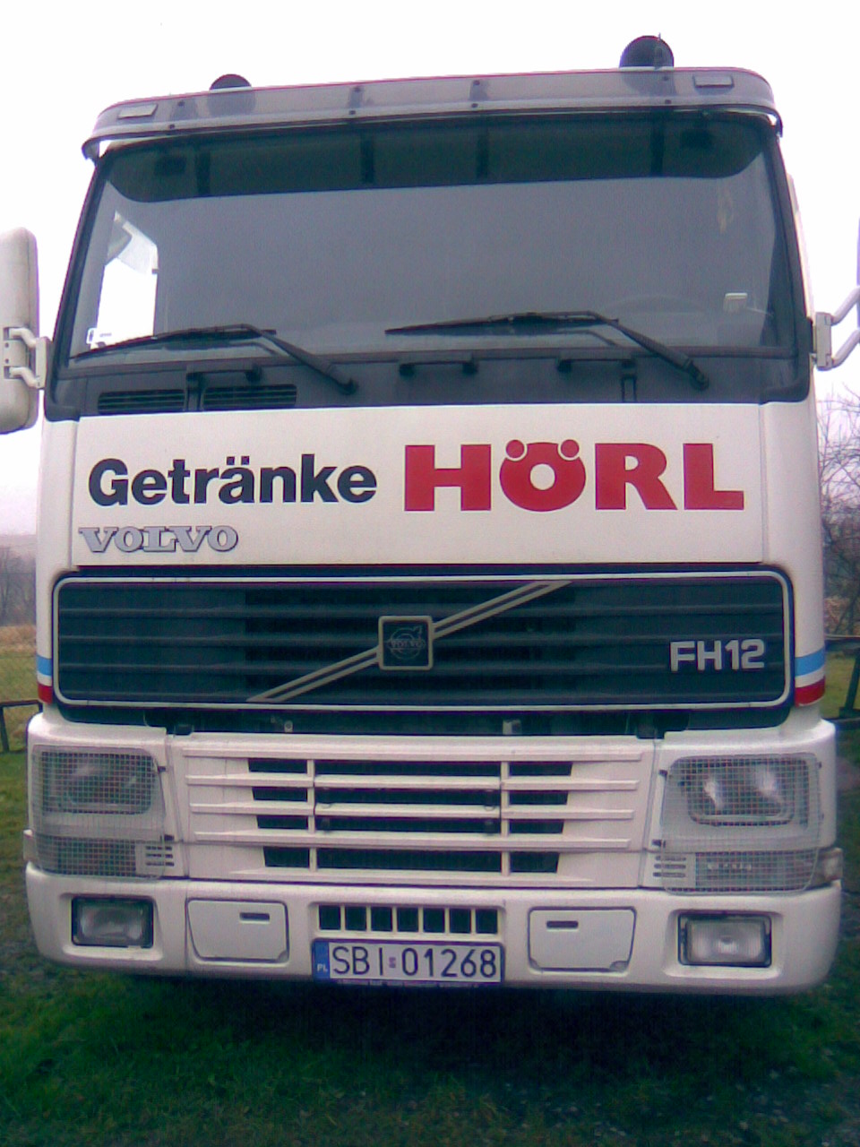 Biała ciężarówka Volvo FH12 z logo 'Getränke HÖRL' na froncie, zaparkowana na trawiastym terenie.