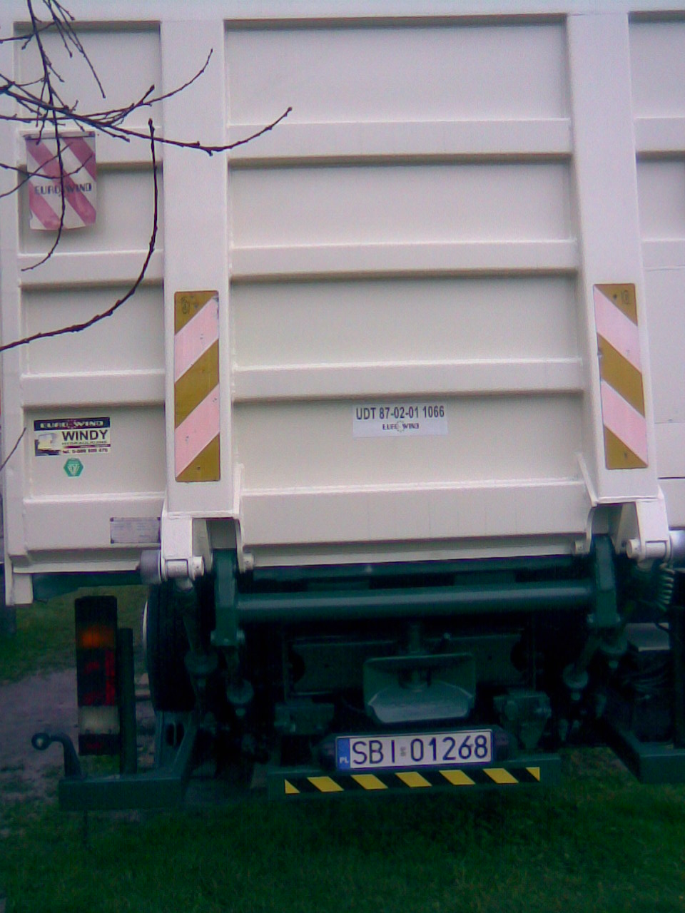 Tył białej ciężarówki z zielonymi elementami podwozia, widoczna tablica rejestracyjna SBI 01268, naklejki ostrzegawcze i gałąź drzewa w lewym górnym rogu.