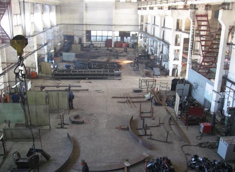 Wnętrze hali produkcyjnej z elementami konstrukcji stalowych w trakcie spawania, widoczny suwnicowy dźwig, spawarka i liczne metalowe elementy.