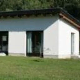 Biały, parterowy dom jednorodzinny z ciemnym dachem, otoczony zielonym trawnikiem. Widoczne duże okna i drzwi przesuwne. Prosta bryła budynku.