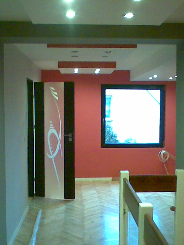 Pomarańczowa ściana w odnowionym pokoju z parkietem, ozdobnymi drzwiami z grafiką i sufitem podwieszanym z oświetleniem punktowym.