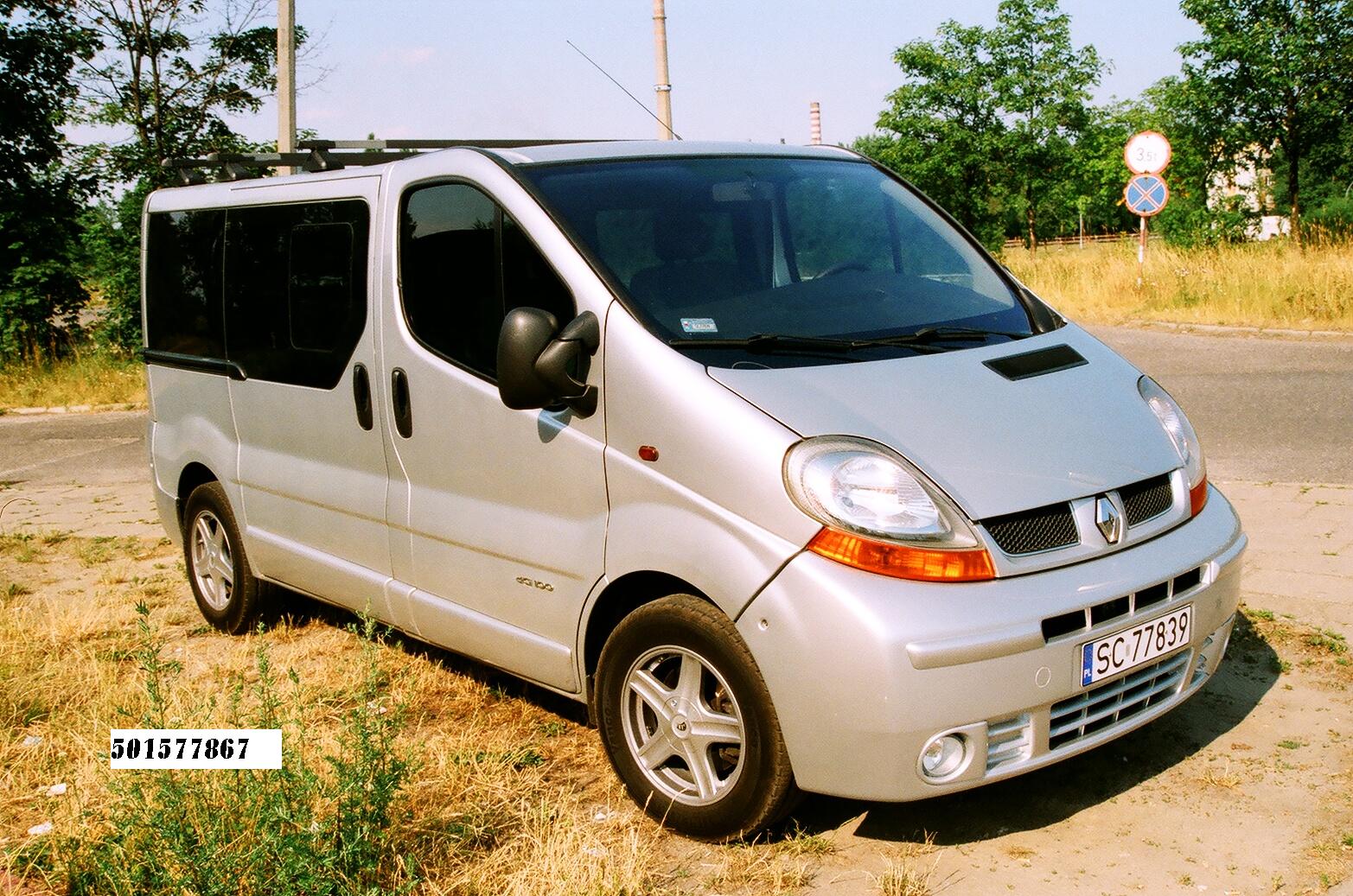 Srebrny bus Renault Trafic z relingami dachowymi, zaparkowany na poboczu z widoczną tablicą rejestracyjną SC 77839 i znakiem drogowym w tle.