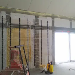 Wnętrze w trakcie prac wykończeniowych: ściany z instalacją ogrzewania podtynkowego, widoczna maszyna do tynkowania i drabina rusztowania.