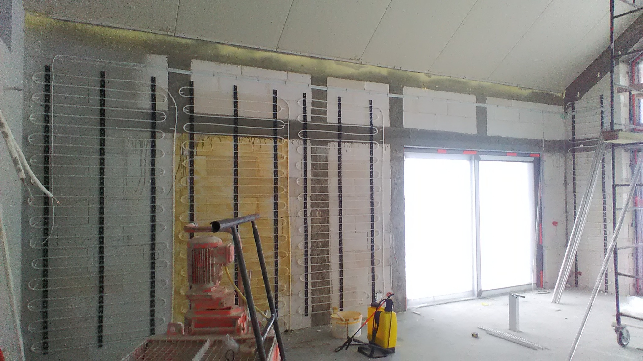 Wnętrze w trakcie prac wykończeniowych: ściany z instalacją ogrzewania podtynkowego, widoczna maszyna do tynkowania i drabina rusztowania.