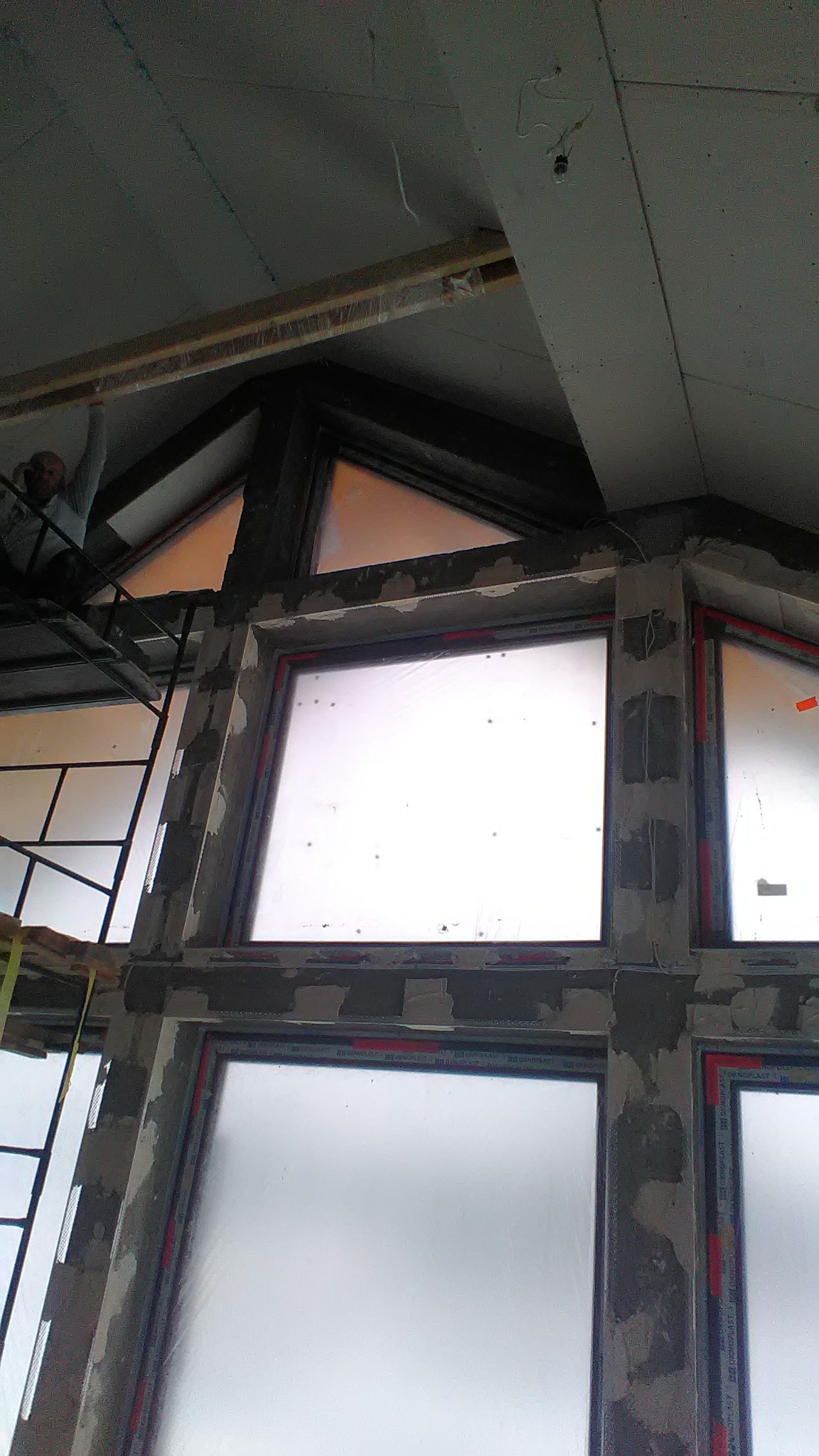 Montaż okien w nowym budynku, widoczne rusztowanie i pracownik na wysokości, surowy stan wykończenia wnętrza.