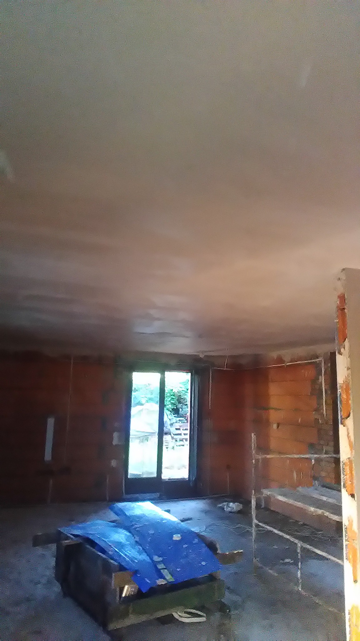 Wnętrze w trakcie prac wykończeniowych: sufit po nałożeniu tynku, ściany z cegły kratówki, okno balkonowe z widokiem na zieleń, rusztowanie i skrzynia z niebieską folią na podłodze.