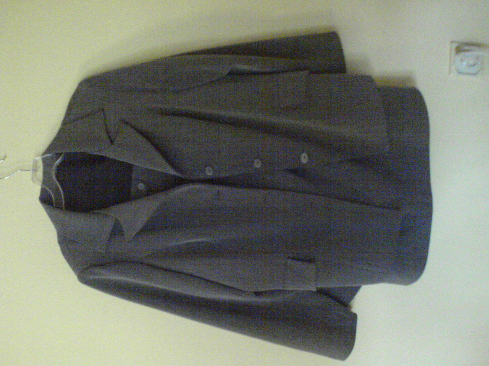 Szary męski blezer z trzema guzikami, wiszący na wieszaku na tle jasnej ściany.