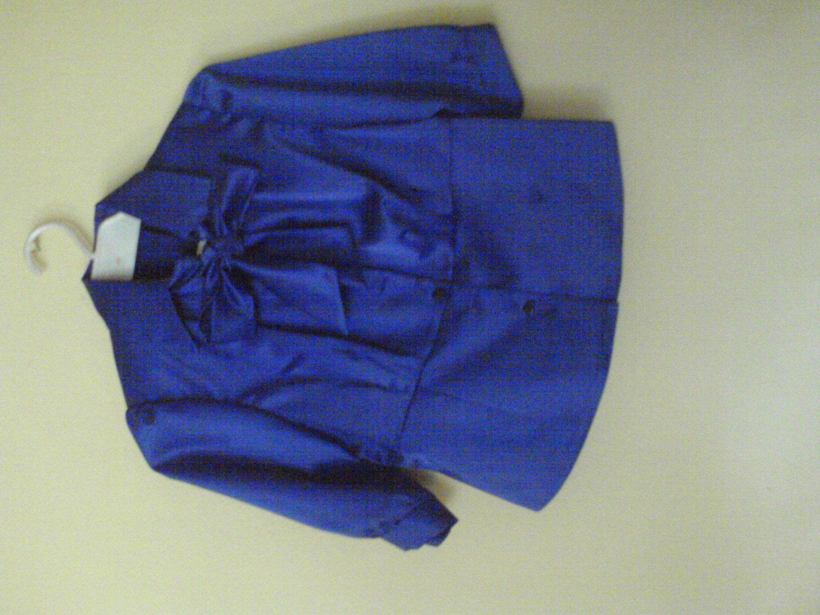 Kobaltowa bluzka damska z kokardą pod szyją, wisząca na białym wieszaku na jasnym tle.