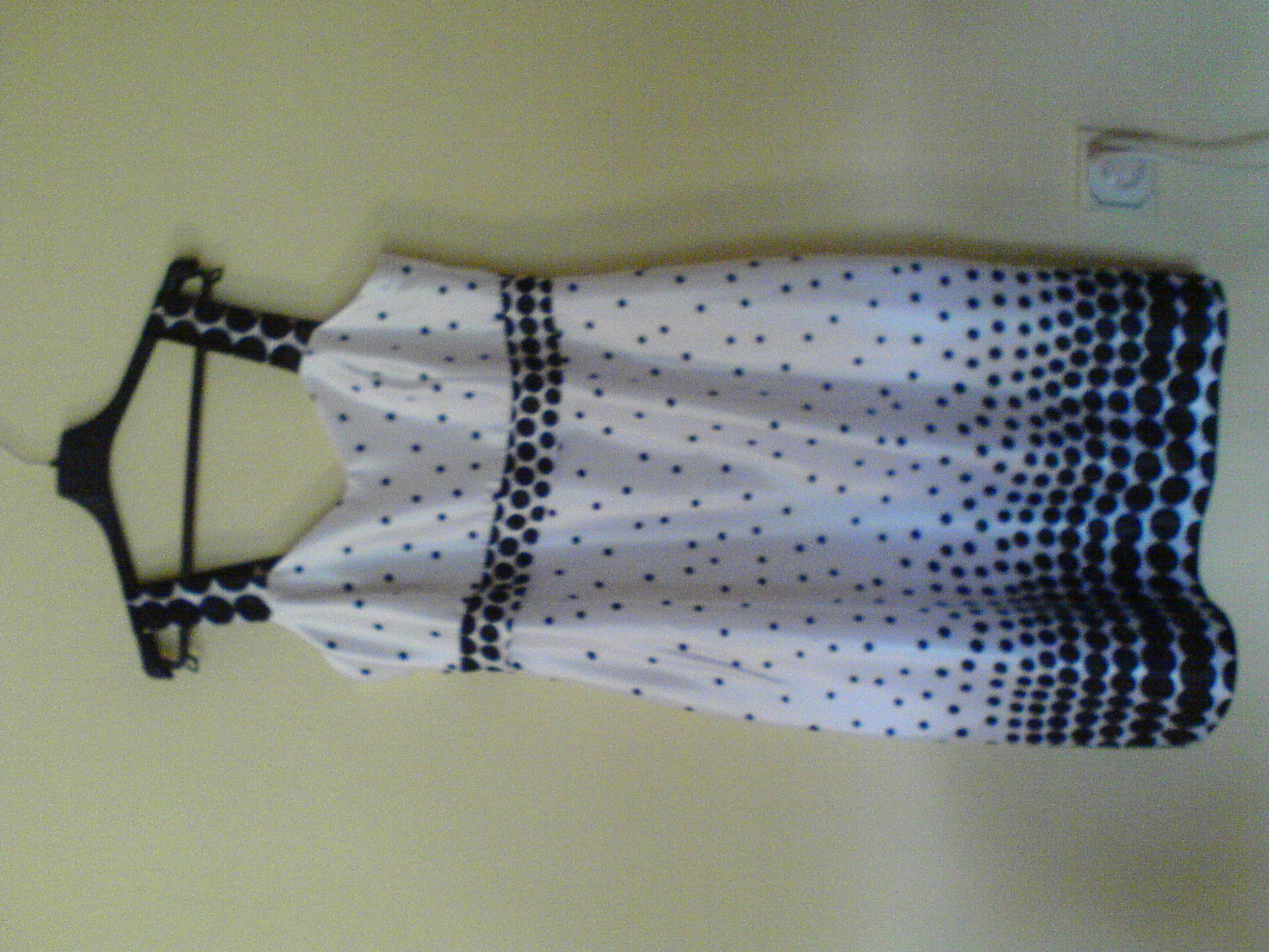 Letnia sukienka w biało-granatowe kropki, wisząca na czarnym wieszaku na tle jasnej ściany. Ujęcie z bliska, widoczne detale wzoru.