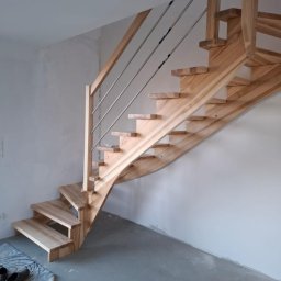 Usługi Stolarskie Walter Patryk - Naturalne schody z jasnego drewna z metalową balustradą, widoczne od dołu, z parą butów na wycieraczce u podstawy. Minimalistyczny design, surowe wykończenie ścian.