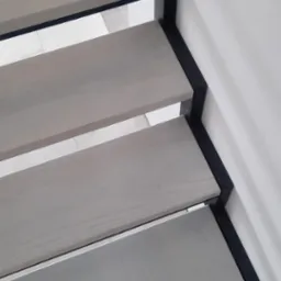 Nowoczesne schody z drewnianymi stopniami w odcieniach szarości i czarną, metalową balustradą. Widok z góry, fragment buta w kadrze. Minimalistyczny design.