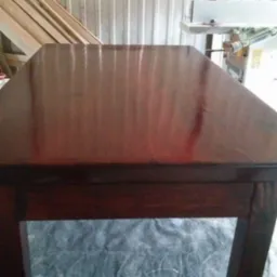 Lakierowany blat drewnianego stołu warsztatowego z widocznymi deskami w tle. Ciemne drewno, odbijające światło, sugeruje świeże wykończenie.