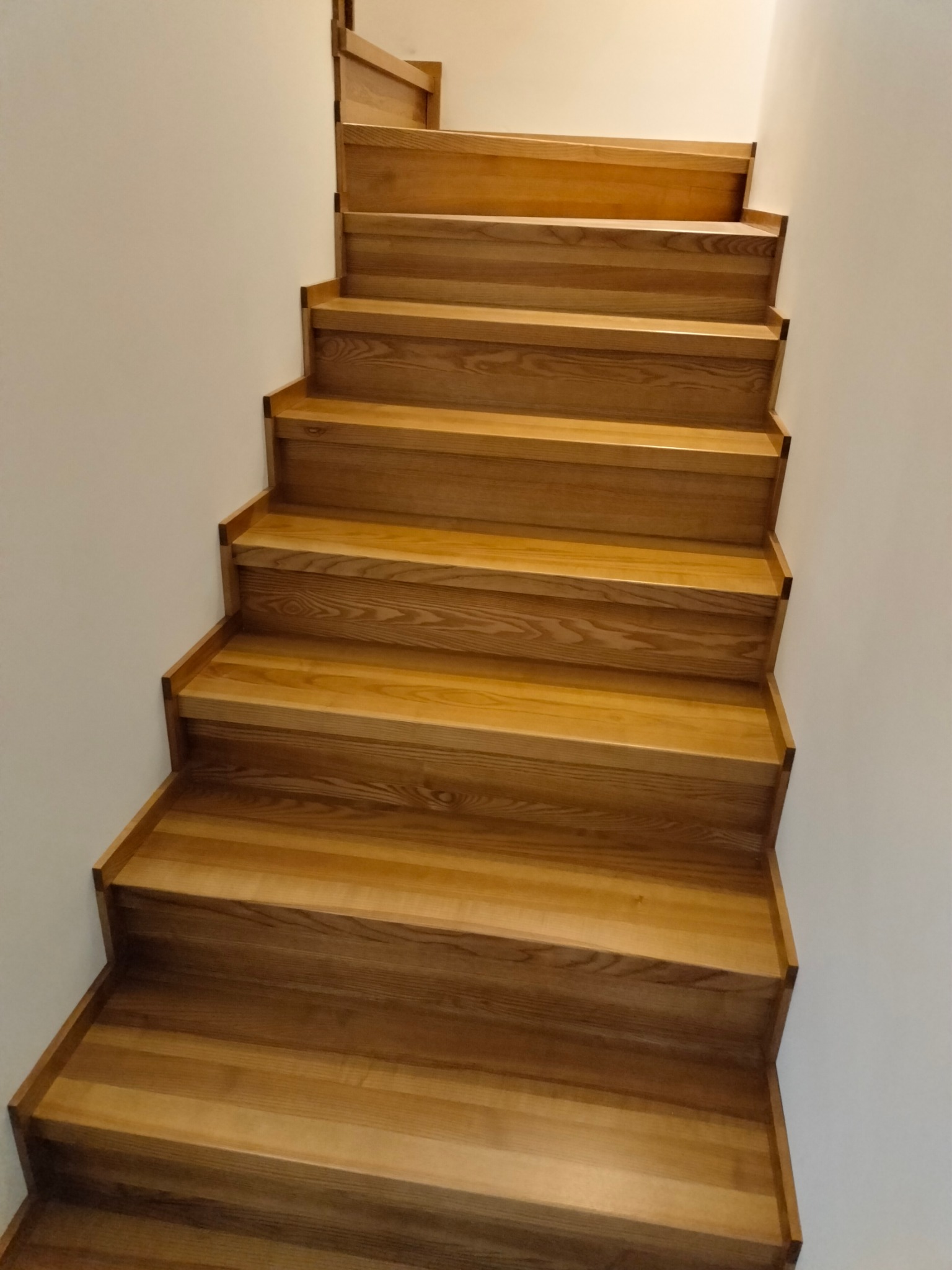 Naturalne, jasne schody drewniane bez poręczy, prowadzące w górę, przylegające do białej ściany. Widoczny detal usłojenia drewna i minimalistyczne wykończenie.