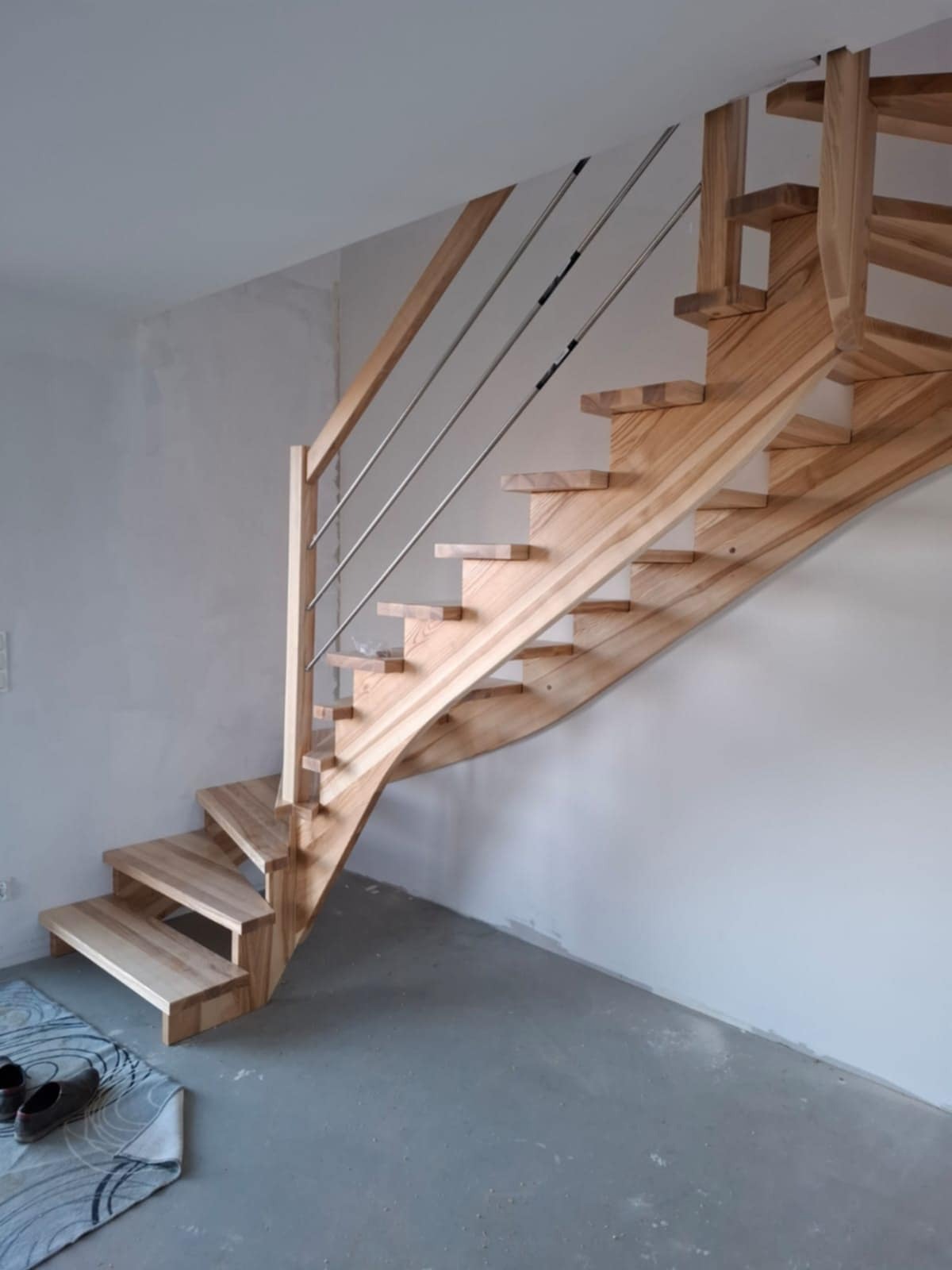 Naturalne schody z jasnego drewna z metalową balustradą, widoczne od dołu, z parą butów na wycieraczce u podstawy. Minimalistyczny design, surowe wykończenie ścian.