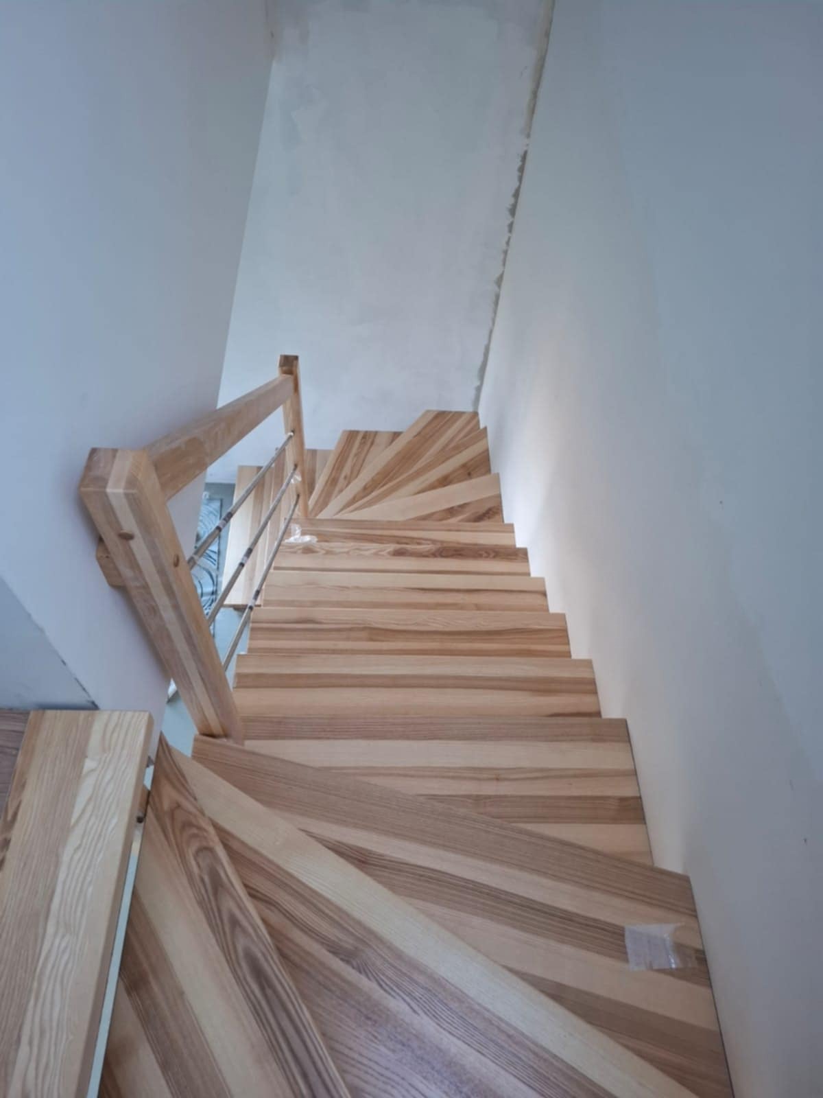 Spiralne schody drewniane z jasnego drewna z metalową balustradą, widziane z góry, w surowym wnętrzu z niepomalowanymi ścianami.