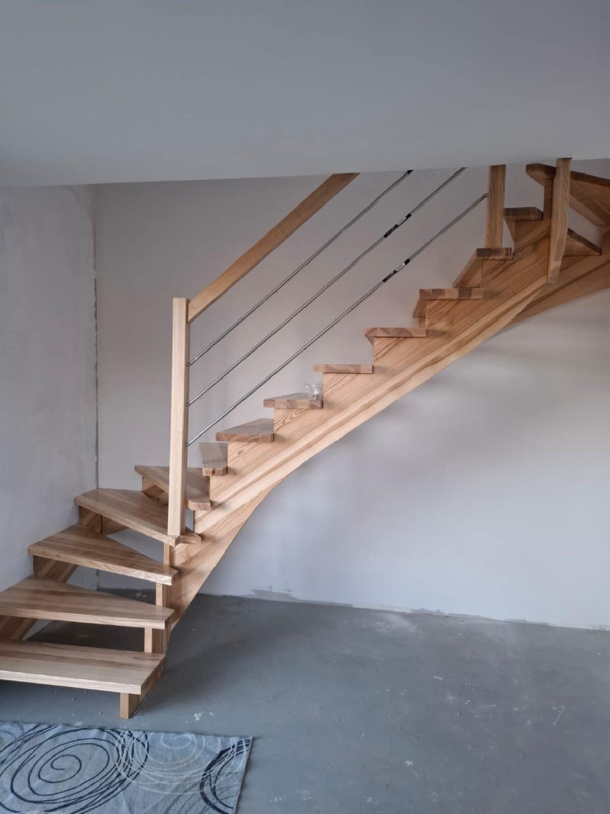 Kręte schody z jasnego drewna z metalową balustradą, widoczne na tle szarej ściany i podłogi. U dołu kadru leży wzorzysty dywanik.