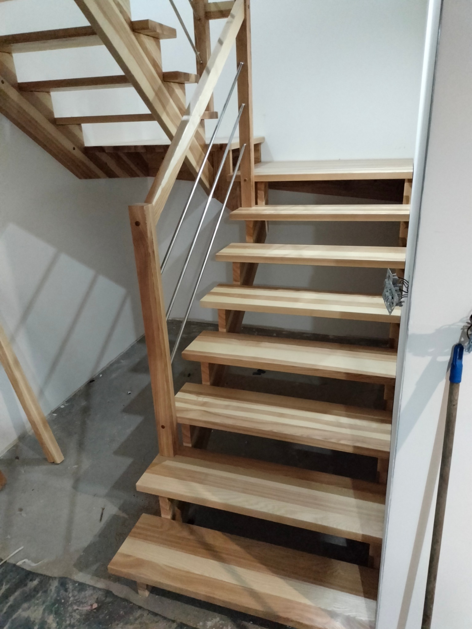 Jasne, drewniane schody z metalową balustradą, widok z dołu. Widoczna surowa podłoga i wystające kable elektryczne, co sugeruje etap wykończeniowy.