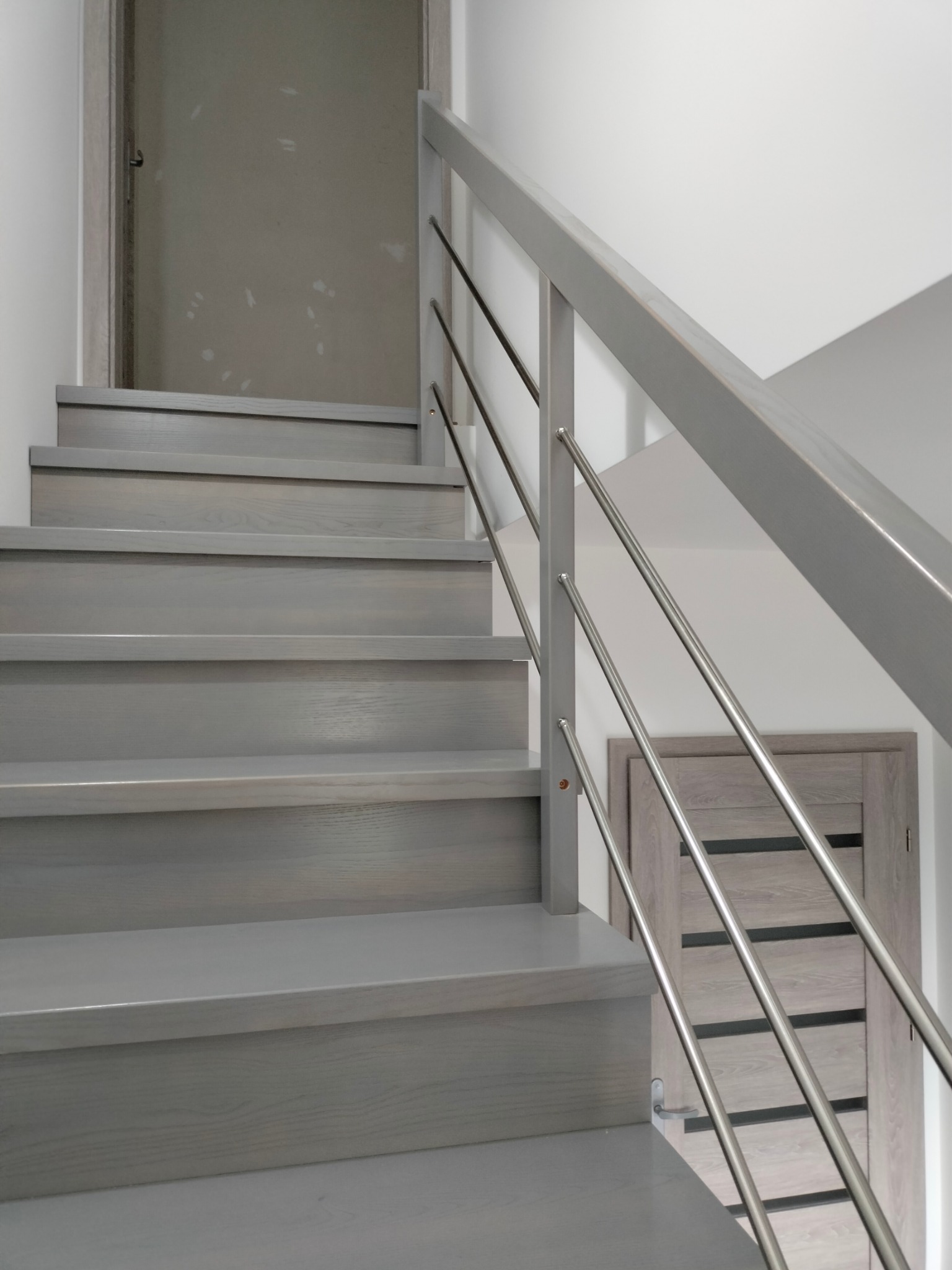 Nowoczesne schody z szarego drewna z metalową balustradą, widok z dołu, minimalistyczny design i jasne wnętrze.