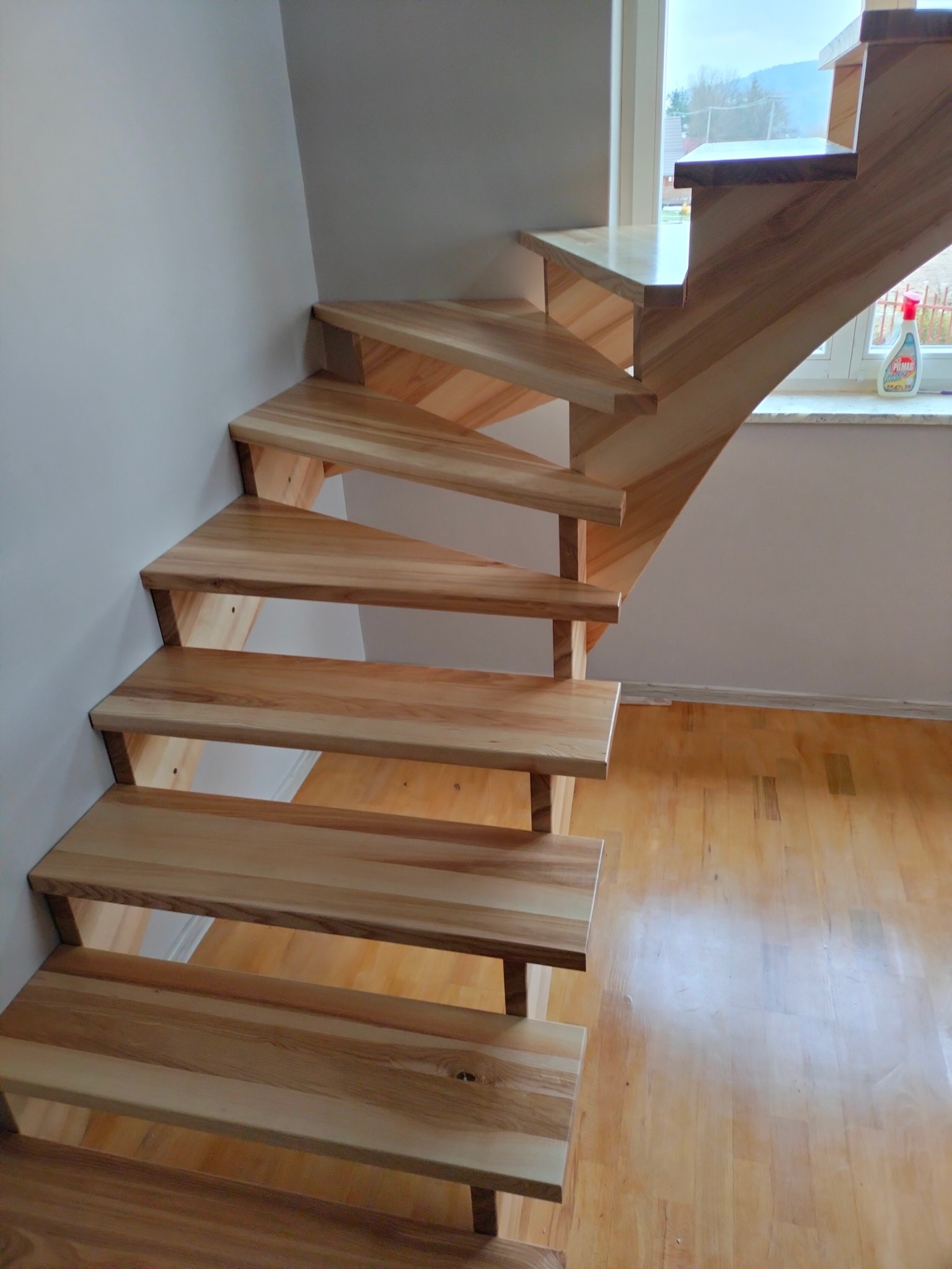 Jasne, drewniane schody bez balustrady, z widoczną strukturą drewna, prowadzące w górę. W tle okno z widokiem na zewnątrz i butelka środka czyszczącego na parapecie.