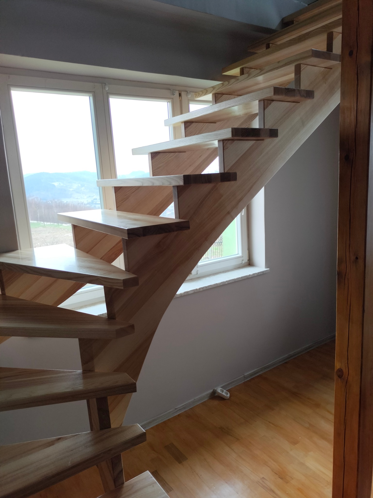 Nowoczesne, drewniane schody samonośne z jasnego drewna, z widokiem na krajobraz za oknem. Minimalistyczny design, brak poręczy, naturalne oświetlenie.