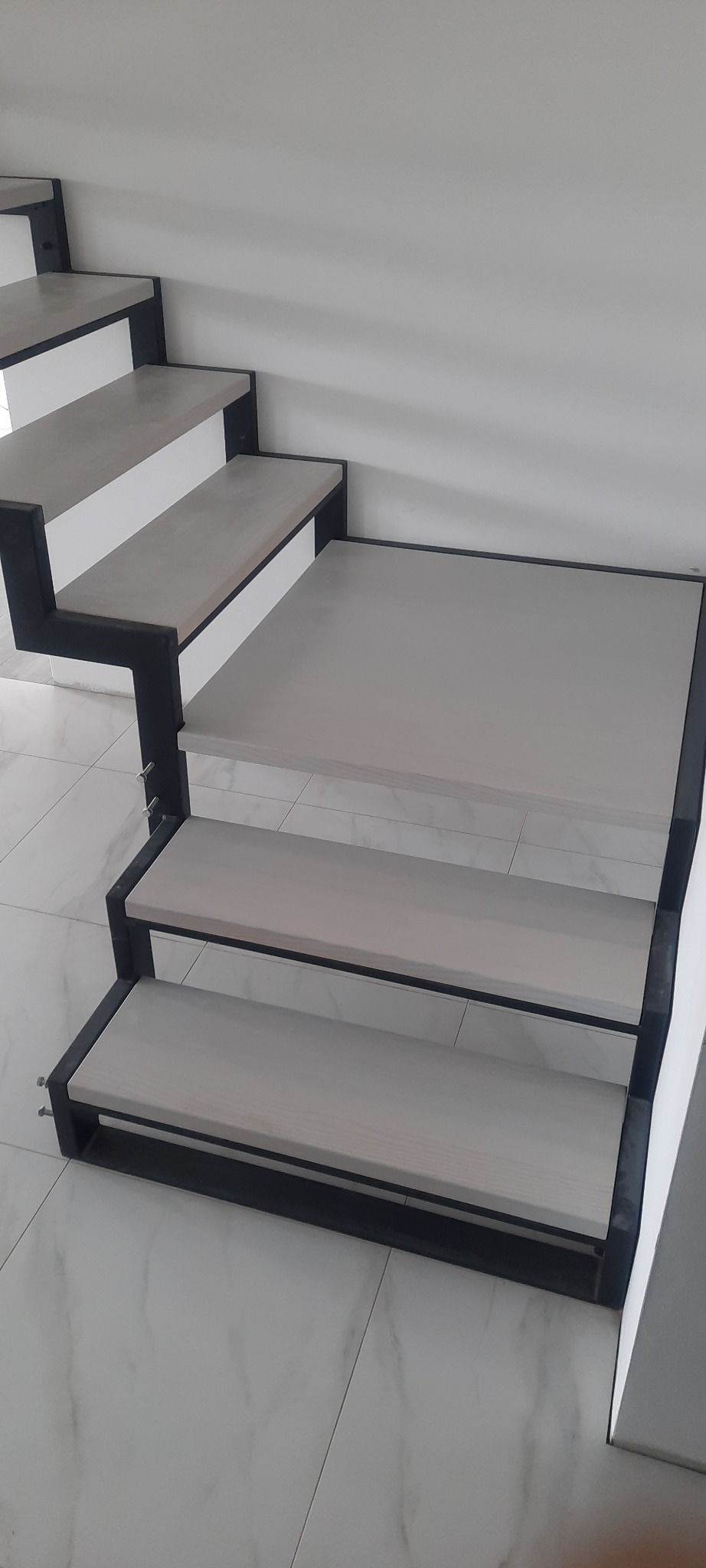 Nowoczesne schody z drewnianymi stopniami w szarym odcieniu i czarną metalową konstrukcją, widok z góry, na tle białej ściany i marmurowej podłogi.