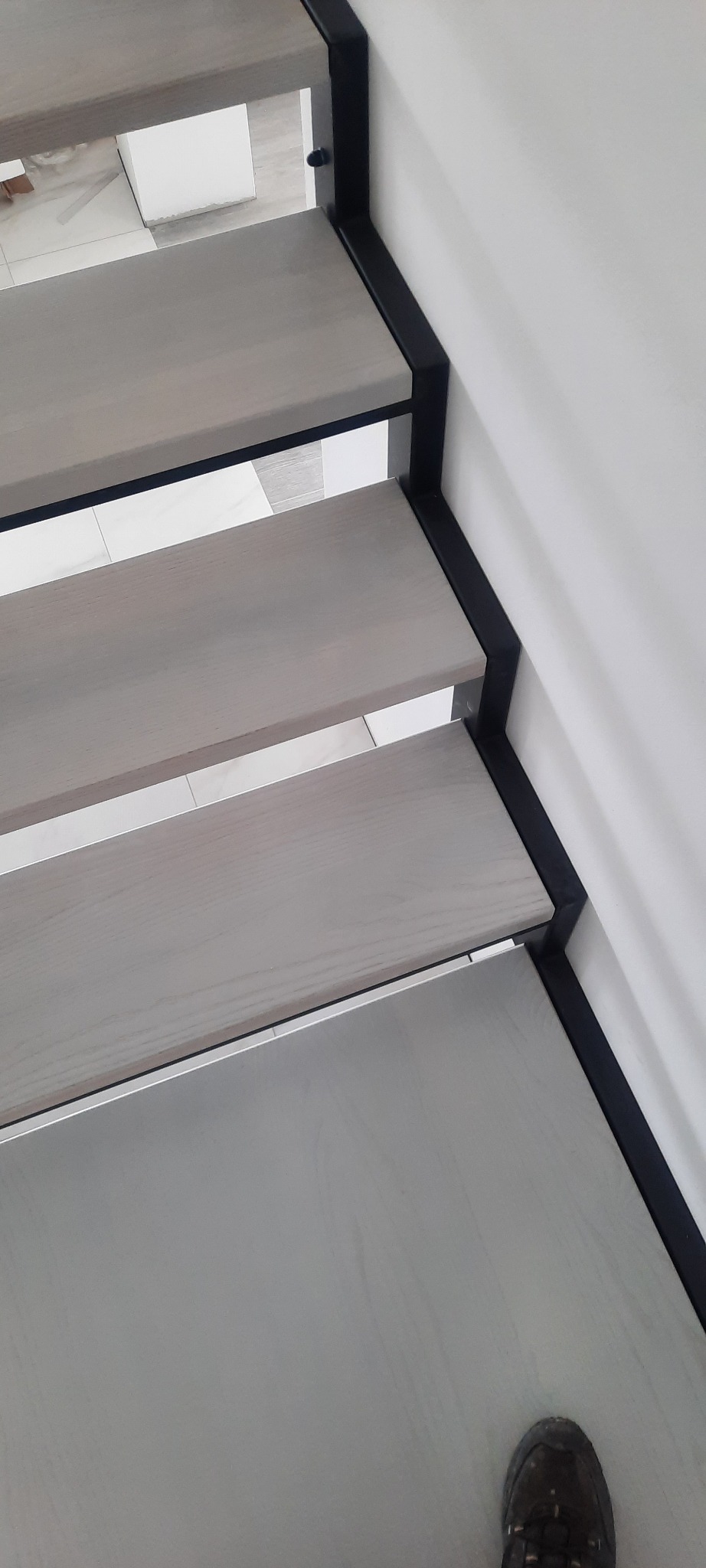 Nowoczesne schody z drewnianymi stopniami w odcieniach szarości i czarną, metalową balustradą. Widok z góry, fragment buta w kadrze. Minimalistyczny design.