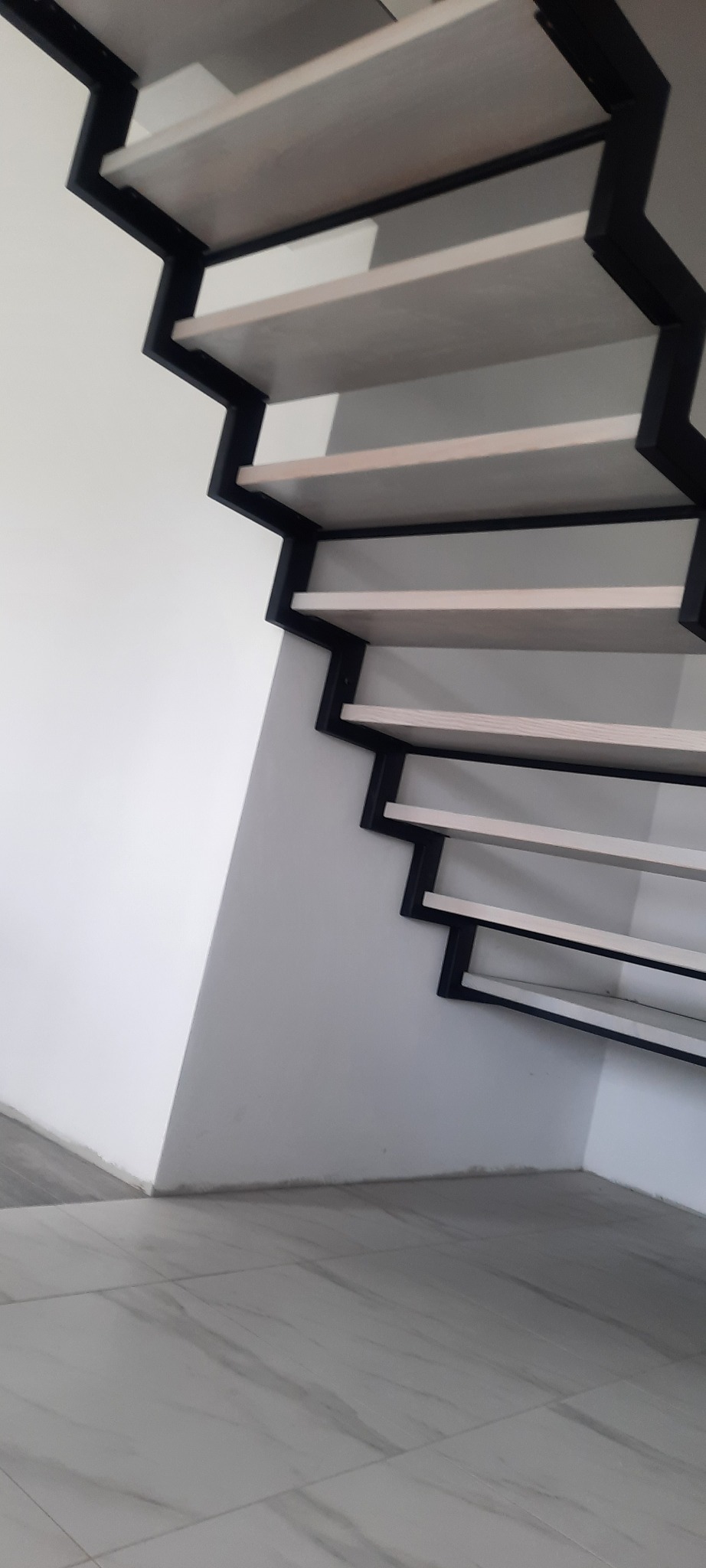 Nowoczesne schody z jasnymi stopniami i czarną metalową konstrukcją, widoczne z dołu, przy białej ścianie i marmurowej podłodze.