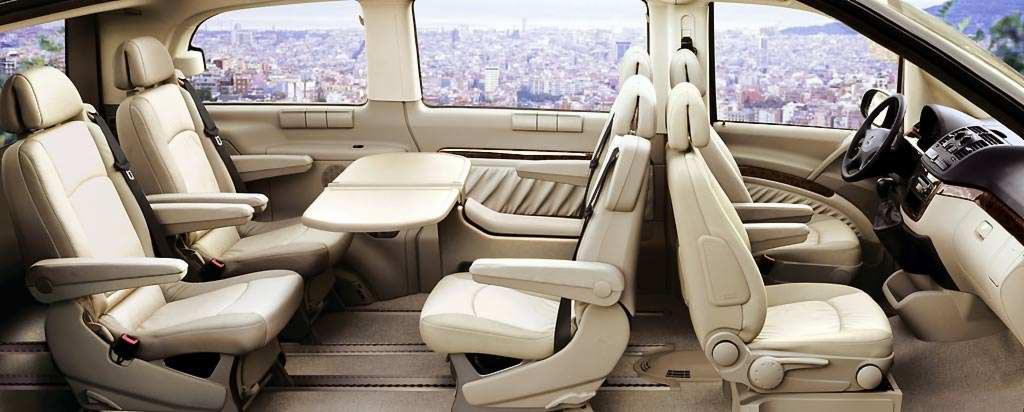 Przestronne wnętrze busa z jasną tapicerką, rozkładanym stolikiem i widokiem na miasto w tle. Komfortowe fotele z regulowanymi podłokietnikami.