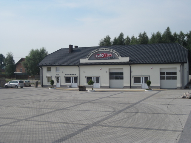 Budynek z napisem 'Regionalne Centrum Kostki Brukowej EUROBRUK plus' z placem wyłożonym szarą kostką, widoczne dwa samochody osobowe i drzewa w tle.