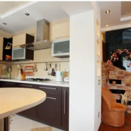 Przestronne wnętrze kuchni z wyspą, brązowymi szafkami i jasnymi blatami, połączone z salonem poprzez dekoracyjną ścianę z cegły i szklaną przegrodę.