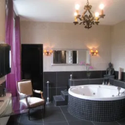 Elegancka łazienka z okrągłą wanną jacuzzi obudowaną czarnymi płytkami, oświetlona ozdobnym żyrandolem i kinkietami, z fioletowymi zasłonami i dekoracyjną figurką Buddy.