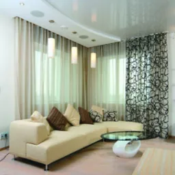 Jasny salon z narożną sofą, szklanym stolikiem kawowym i designerskimi lampami wiszącymi, ujęcie z perspektywy lekko z góry, ukazujące podwieszany sufit z wbudowanym oświetleniem.