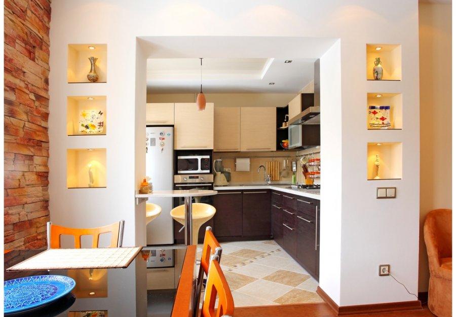 Nowoczesna kuchnia z jadalnią, ściana z cegły dekoracyjnej, wnęki z oświetleniem eksponujące ozdobne przedmioty, widok na zabudowę kuchenną w odcieniach beżu i brązu.