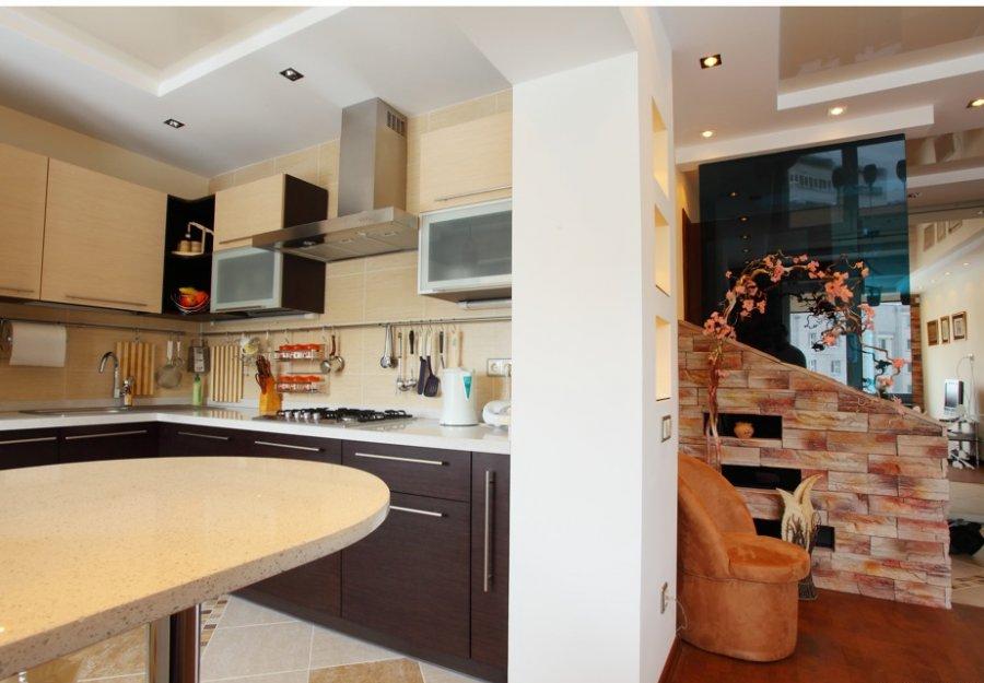 Przestronne wnętrze kuchni z wyspą, brązowymi szafkami i jasnymi blatami, połączone z salonem poprzez dekoracyjną ścianę z cegły i szklaną przegrodę.