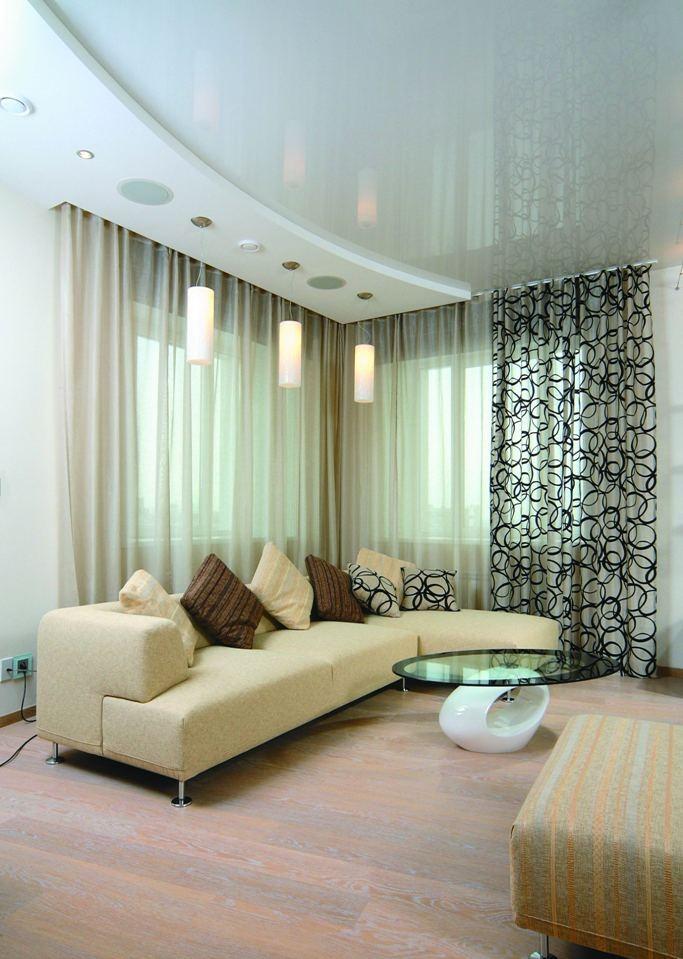 Jasny salon z narożną sofą, szklanym stolikiem kawowym i designerskimi lampami wiszącymi, ujęcie z perspektywy lekko z góry, ukazujące podwieszany sufit z wbudowanym oświetleniem.
