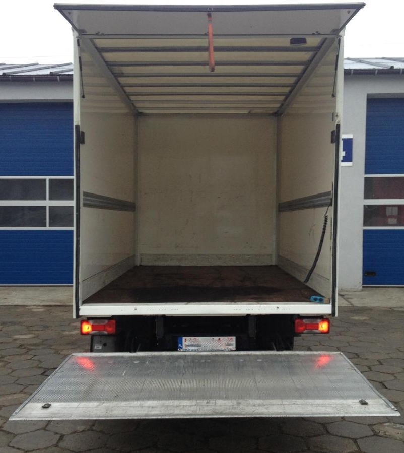 Pusta przestrzeń ładunkowa białego samochodu dostawczego z otwartą klapą, widok z tyłu na tle niebieskich bram garażowych.