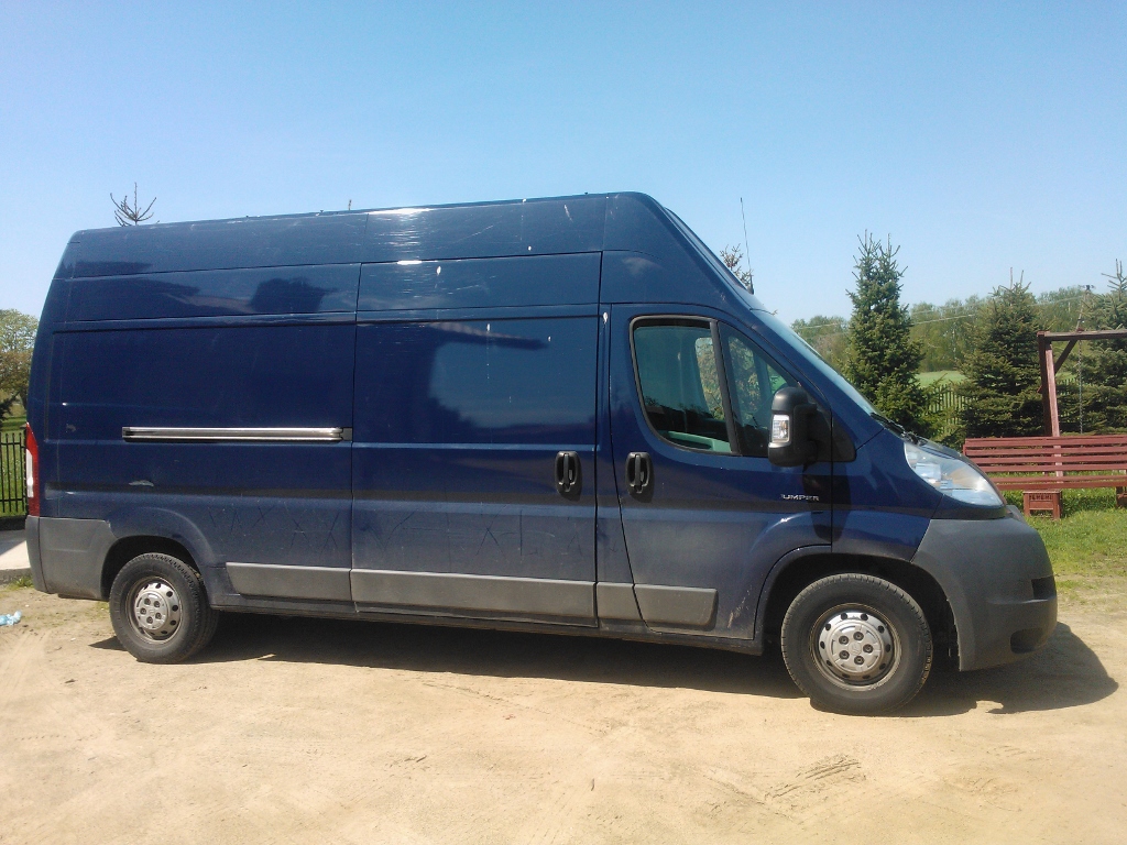 Granatowy, wysoki bus dostawczy marki Citroen Jumper na piaszczystym podłożu, z widocznymi śladami użytkowania na karoserii, sfotografowany w słoneczny dzień na tle zieleni i błękitnego nieba.