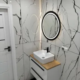 Nowoczesna łazienka z biało-czarnymi płytkami imitującymi marmur, okrągłe lustro z podświetleniem LED, biała umywalka nablatowa na szafce z drewnianym blatem i czarnymi uchwytami.
