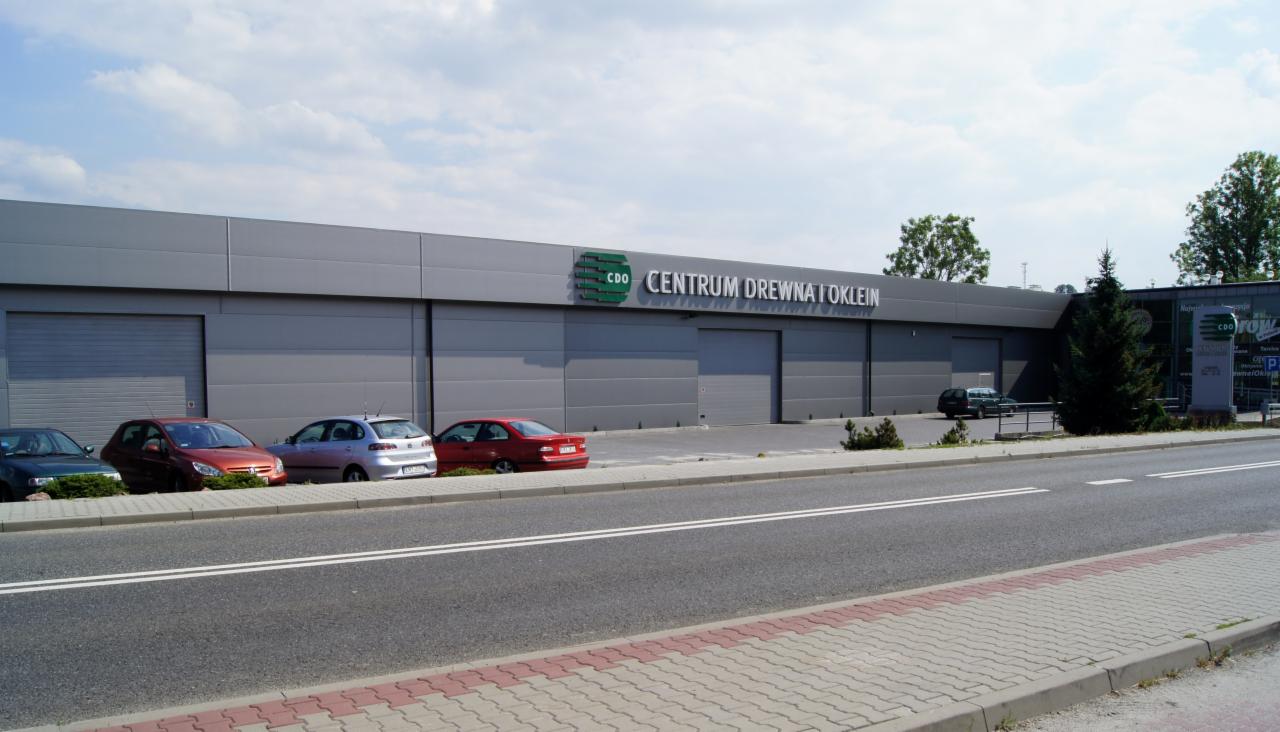 Fasada budynku Centrum Drewna i Oklein z szarymi panelami, widoczne logo firmy i zaparkowane samochody wzdłuż ulicy.