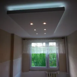 Podwieszany sufit z oświetleniem punktowym w jasnym pomieszczeniu. Widok na okno z koronkową firanką i grzejnik. Ściany w beżowym odcieniu.