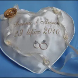 Poduszka w kształcie serca z haftowanym napisem 'Iwona i Marek 29 Lipiec 2010', zdobiona wstążkami i różyczkami, z dwoma złotymi obrączkami umieszczonymi na środku.